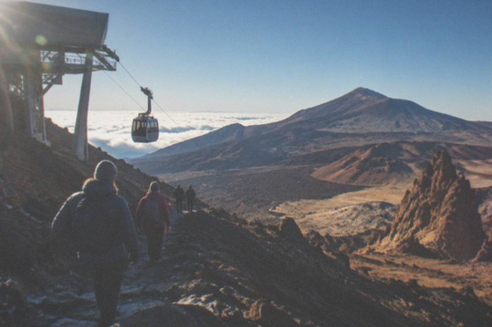 Hikers in warm jackets walking a rocky path below a cable car station