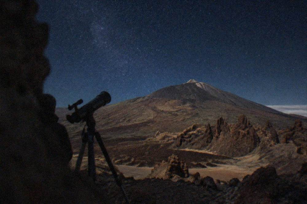 A telescope silhouetted against a starry night sky above a volcanic landscape