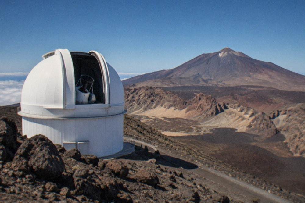 A large white observatory dome with a slit open toward the sky on a rocky volcanic ridge