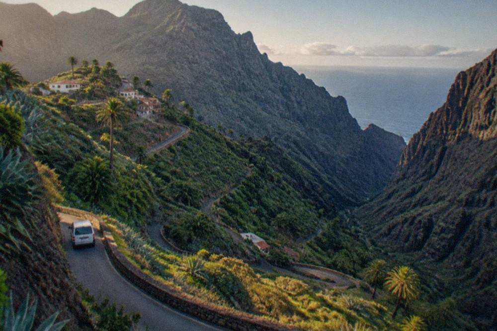 A narrow mountain road winding through green terraced hills towards Masca village