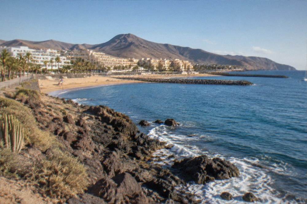 A calm southern Tenerife bay in summer with clear water and a paved seafront behind it