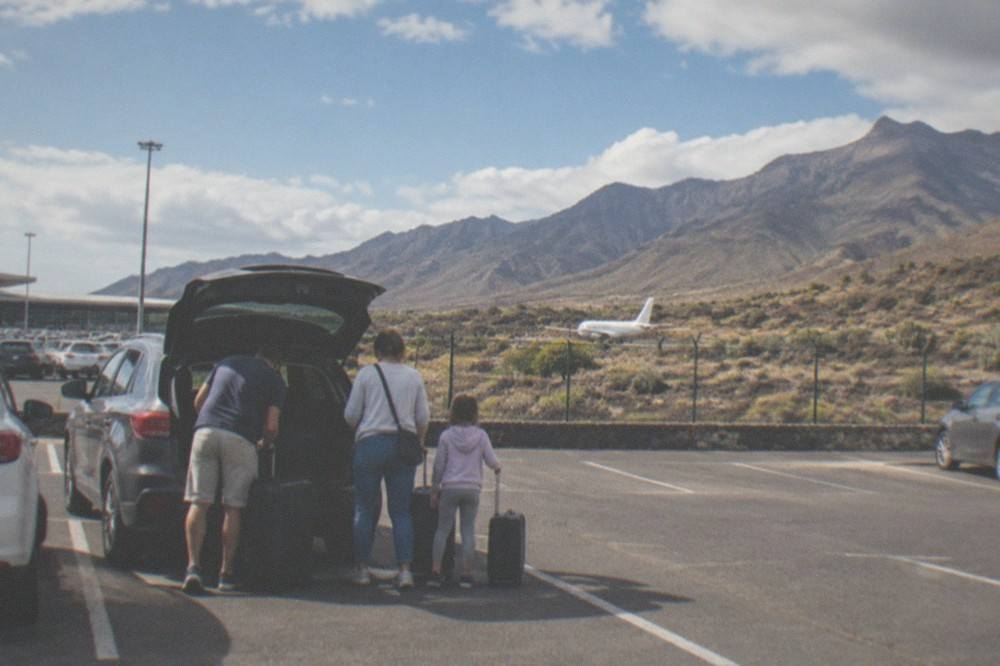 A family loading suitcases into the boot of a car in an airport car park with mountains in the background