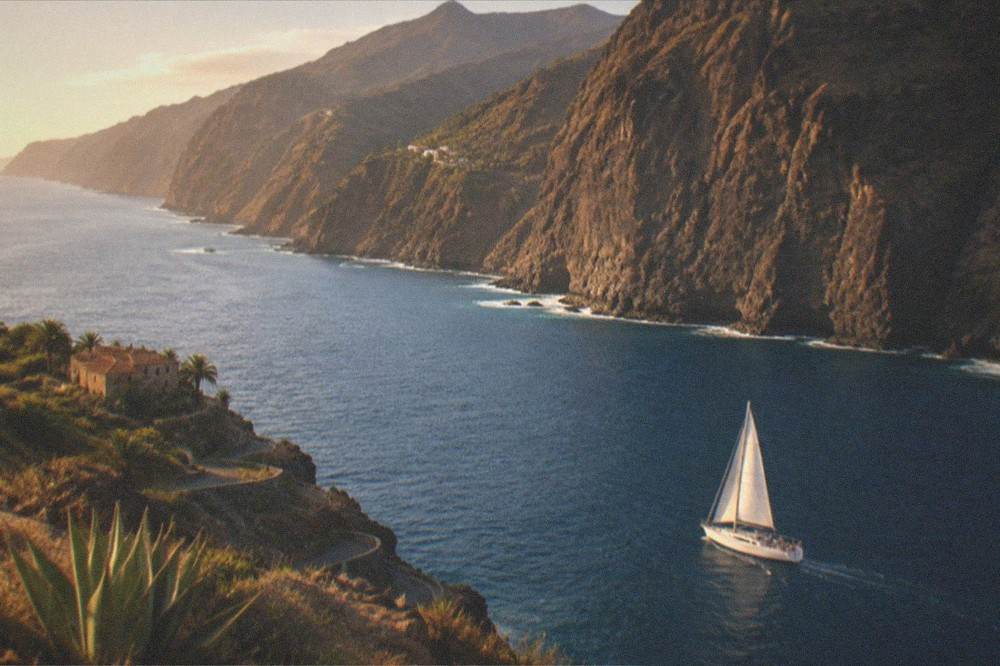 A sailboat under way on calm blue water near the sea cliffs of Los Gigantes with passengers on deck