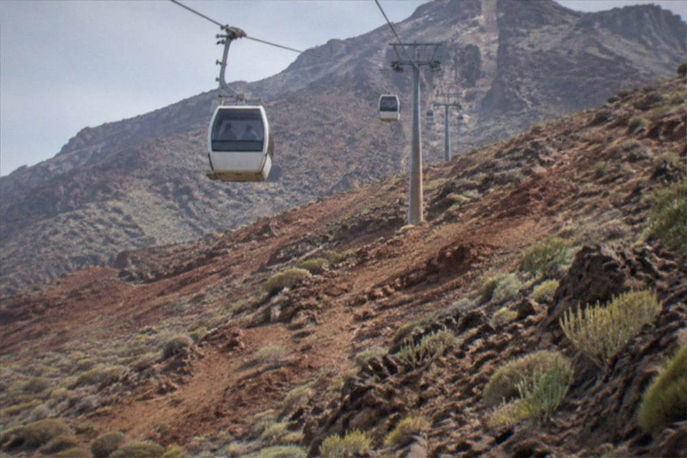 A cable car cabin ascending over a red and grey volcanic mountainside towards high peaks