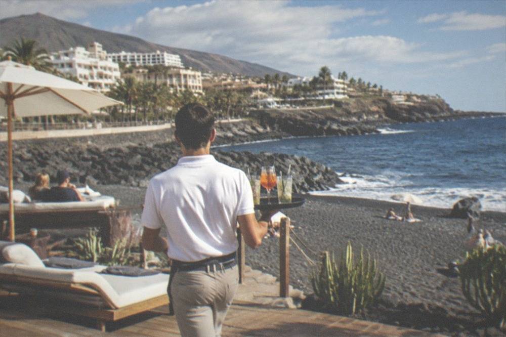 A waiter carrying drinks across a beach club deck towards the sea