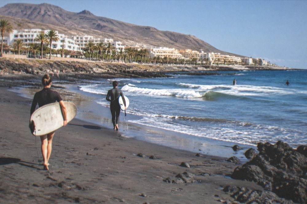 Surfers carrying boards across a beach towards small waves