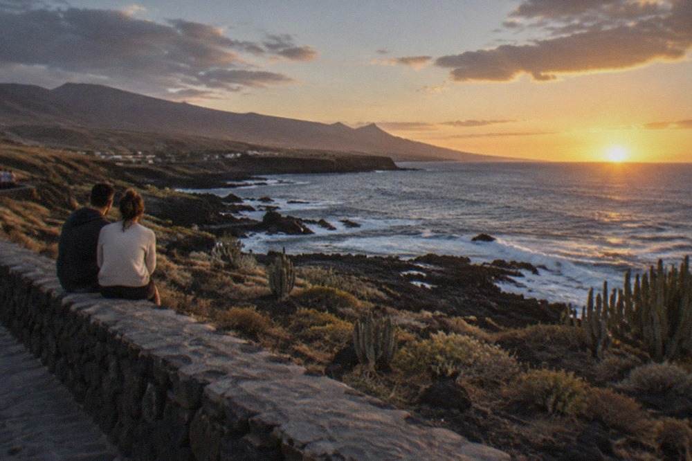 A couple sitting on a sea wall watching the sunset over the Tenerife coastline