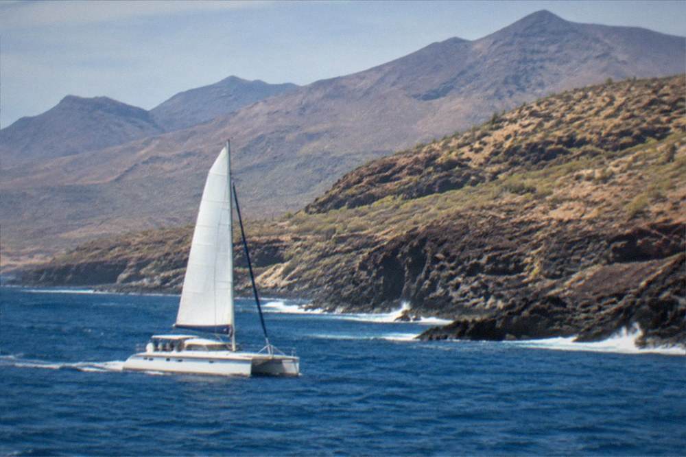 A catamaran sailing past a volcanic coastline with mountains behind