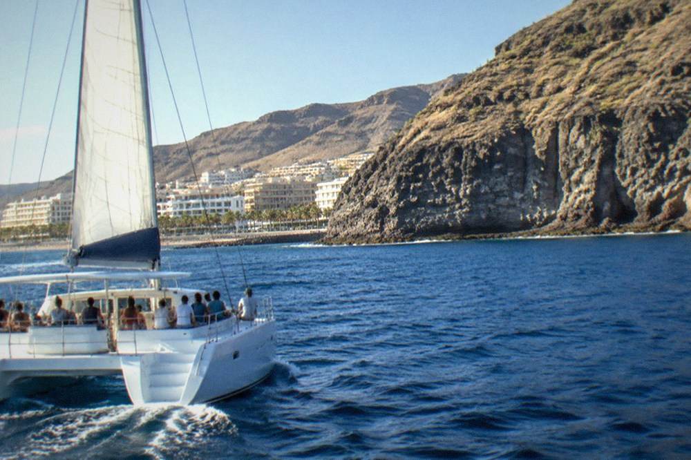 A catamaran with families on deck passing tall sea cliffs on a Tenerife coast
