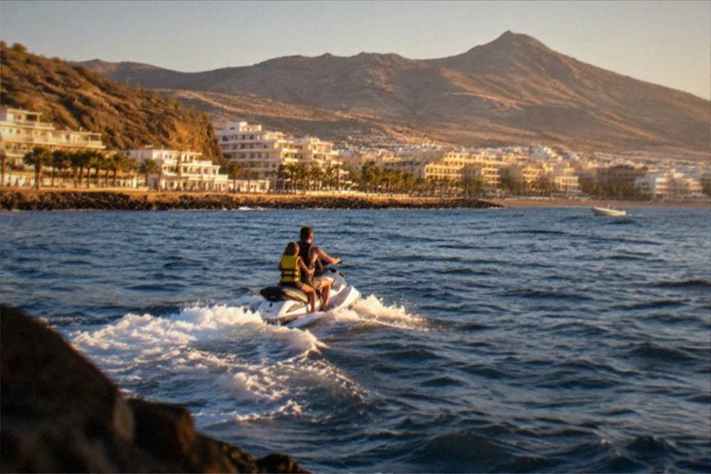 A child in a life jacket riding behind an adult on a jet ski near a sunny coast