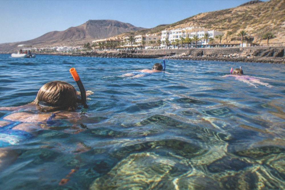 A family with snorkels swimming over clear water above a rocky reef