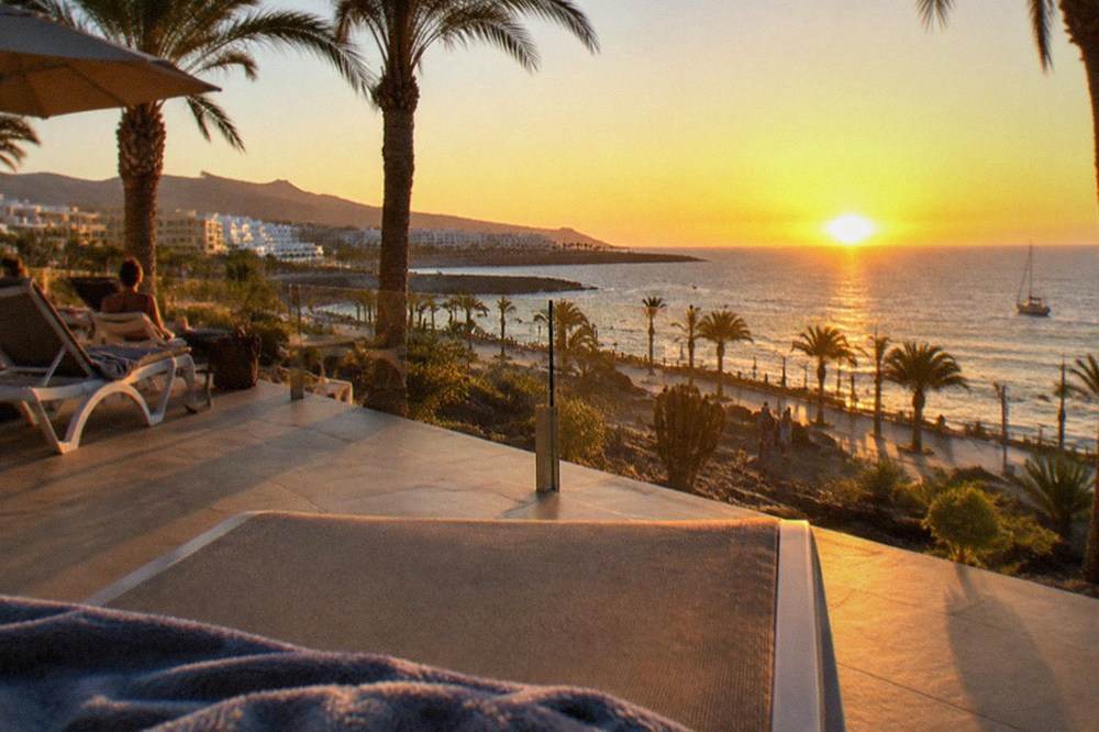 A hotel terrace with palm trees and sun loungers facing the sea at sunset