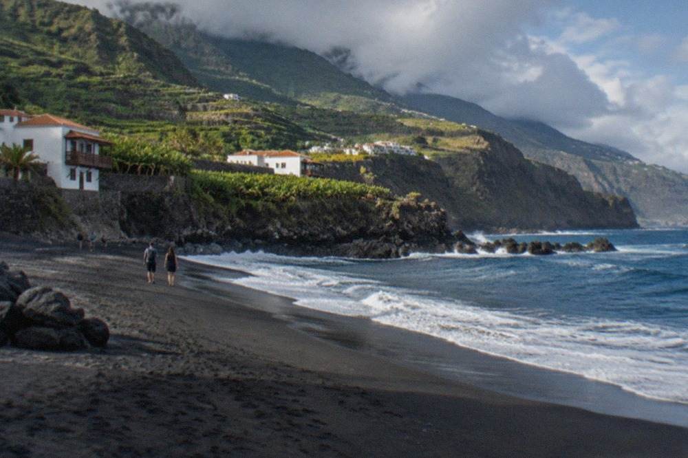 A wide volcanic beach of dark sand with waves breaking under a cliff