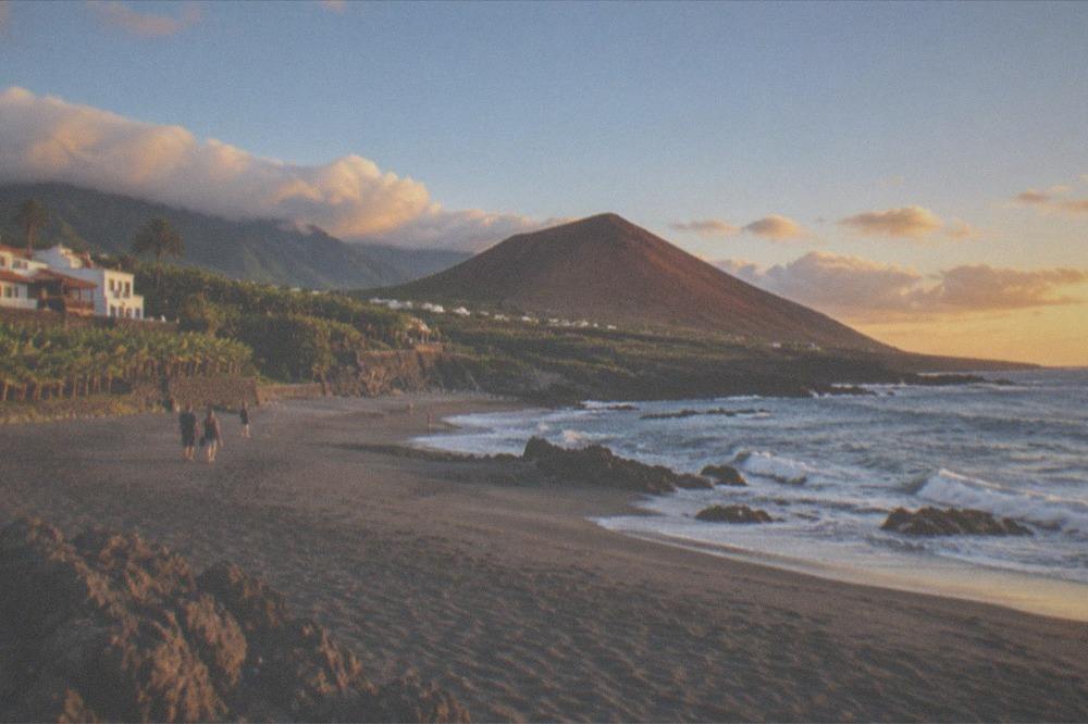 A grey sand beach stretching out beneath a small red volcanic cone
