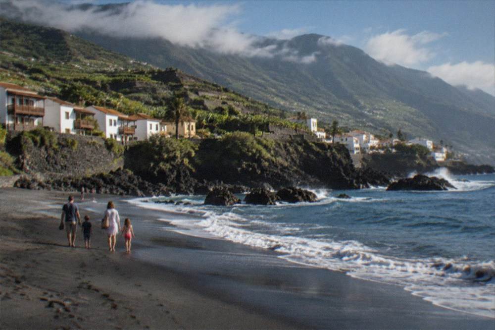 A family walking along a dark sand shoreline in bright morning light