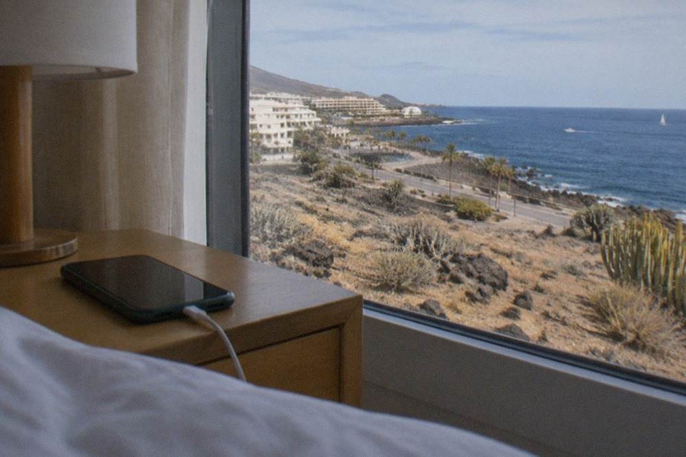 A phone charging on a hotel room bedside table beside a window with a view of the sea