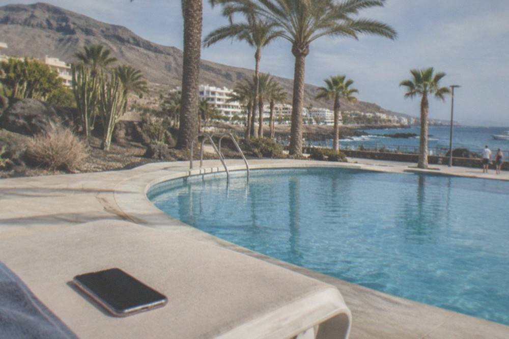 A smartphone resting face up on a sun lounger beside a swimming pool with palm trees