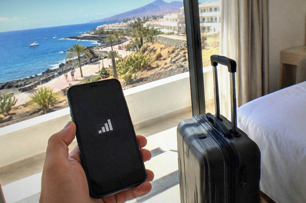 A traveller holding a smartphone showing a network signal beside a suitcase in a bright hotel room