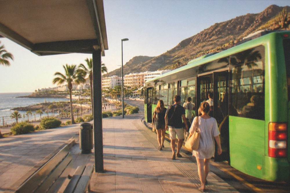 A bus station platform with a green island bus boarding passengers under a sunny sky