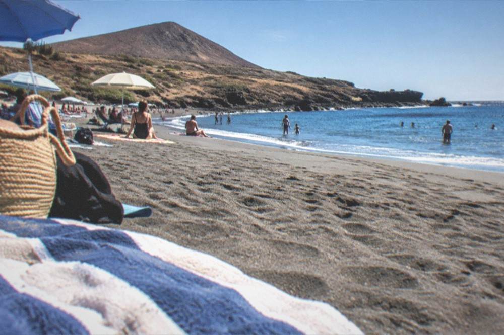 A crowded beach of dark sand at El Medano with sun umbrellas and calm water in winter sunshine