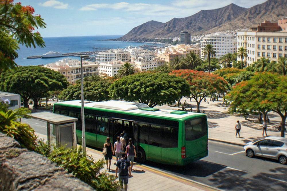 Passengers boarding a green island bus at a stop in a sunny resort town
