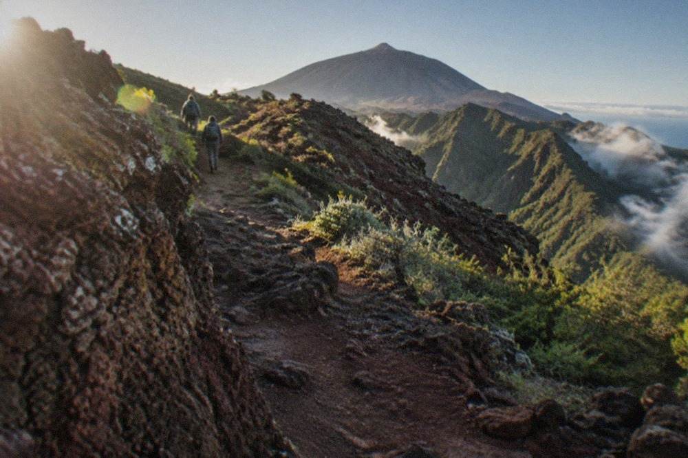 A rocky volcanic footpath climbing through red and ochre lava fields toward Mount Teide under a clear blue sky
