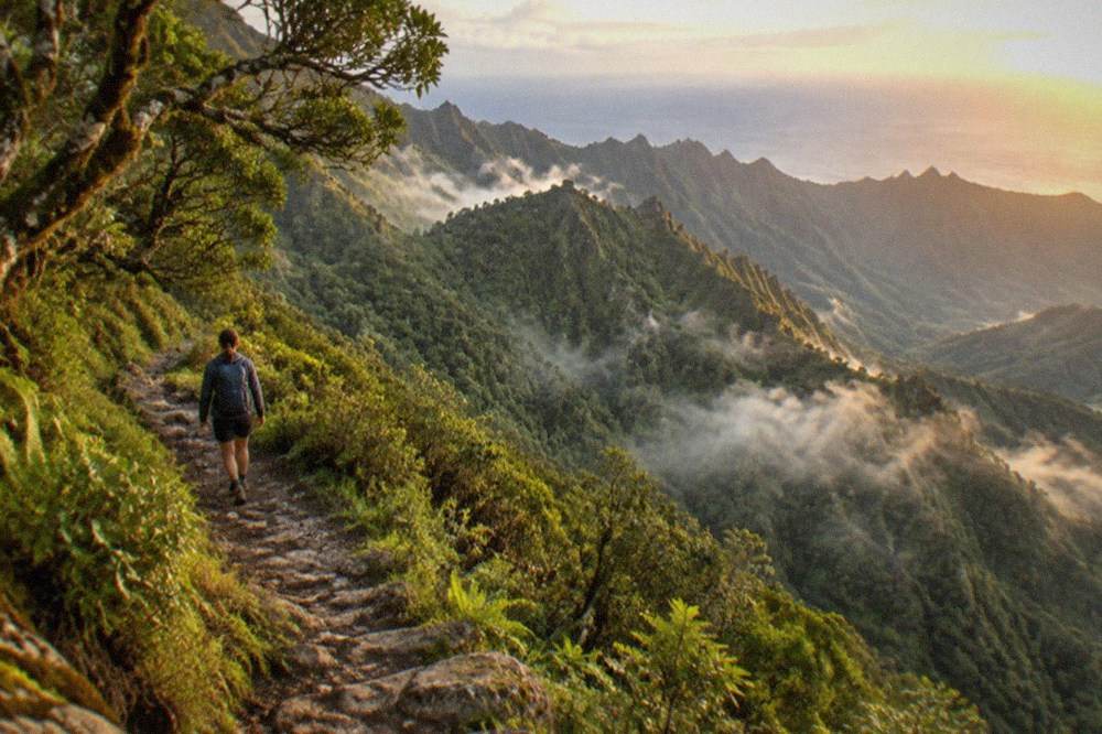 A hiker with a backpack walking a narrow stony path above a deep green ravine in the Teno mountains