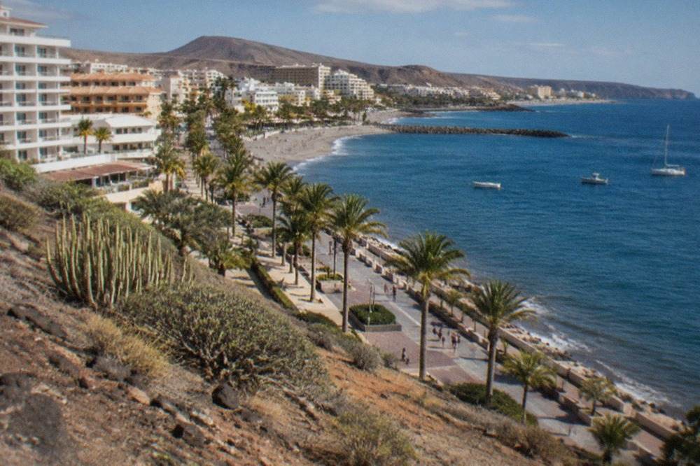 A view along a sunny Tenerife south coast promenade with palm trees, hotels and calm blue sea