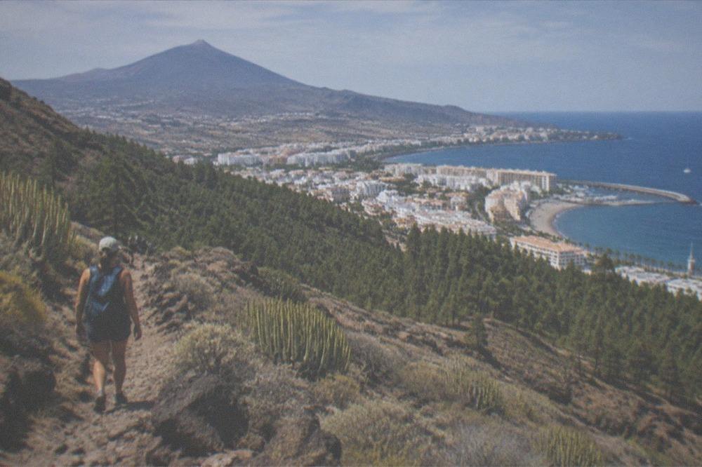 A hiker on a rocky mountain trail above pine forest with a volcano in the distance