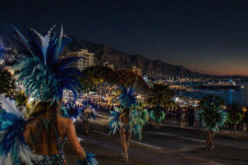 Dancers in feathered carnival costumes performing in a night-time parade under stage lights