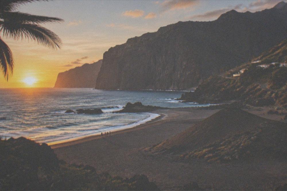 A wide grey-sand beach with a small volcanic cone at dusk