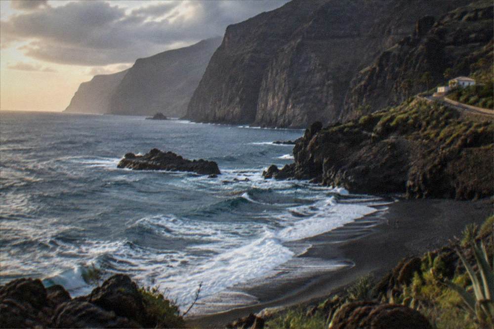 A black sand cove beneath a cliff with waves in fading light