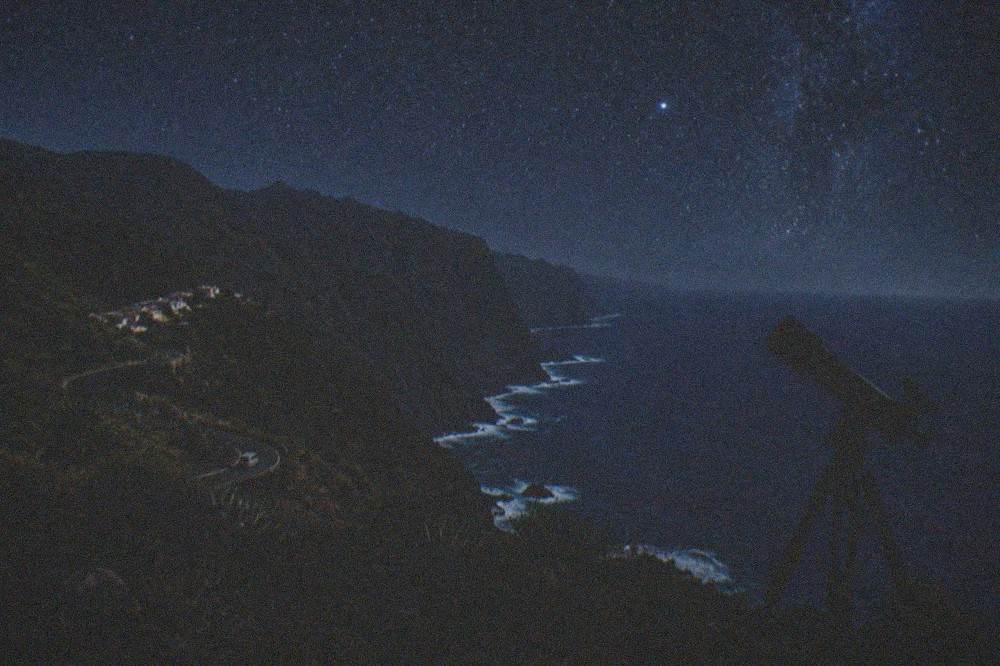 A telescope silhouetted against a starry night sky above dark volcanic ground