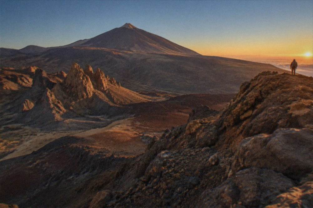 Sunrise light on the summit cone seen from a high ridge with a hiker silhouette in the distance