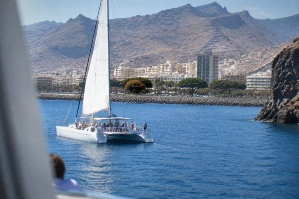 A catamaran on calm blue water near tall sea cliffs with people watching from the deck
