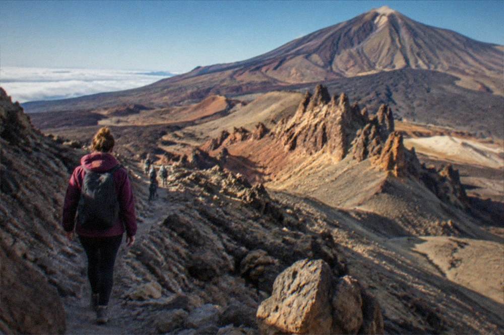 A hiker in a warm layer walking a rocky volcanic trail above the clouds on Teide