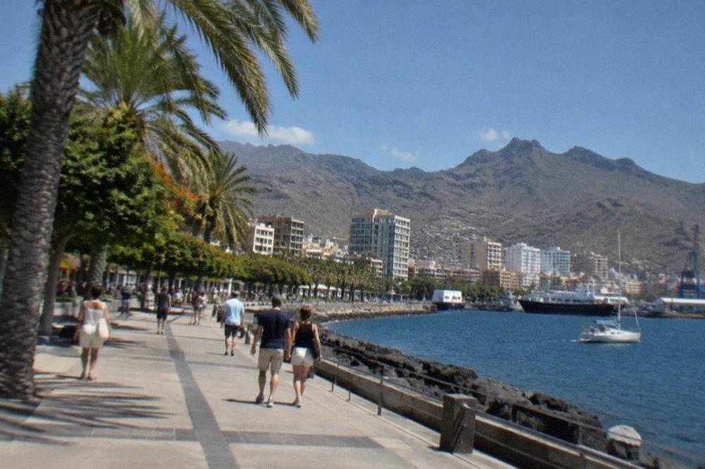 A seafront promenade in Santa Cruz de Tenerife with people in light clothing walking under palm trees