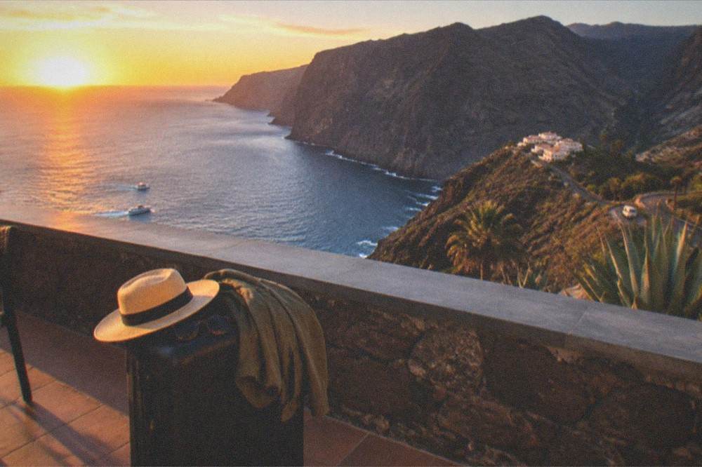 A suitcase with a sun hat, sunglasses and a folded jacket on a terrace overlooking the sea at Los Gigantes at sunset