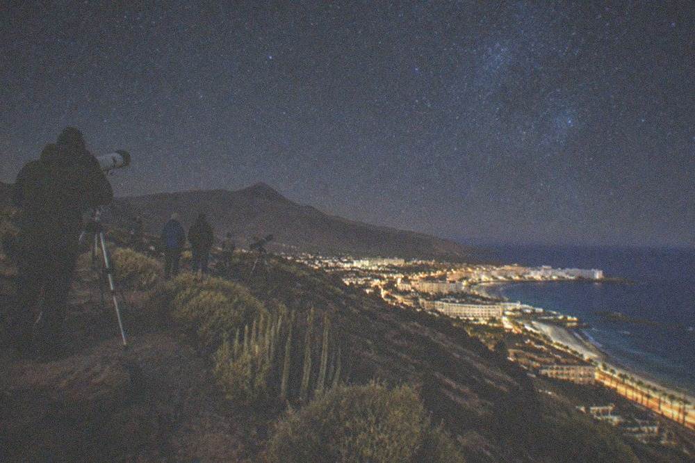 Telescopes and people in warm coats under a star-filled sky on a mountain ridge in Tenerife