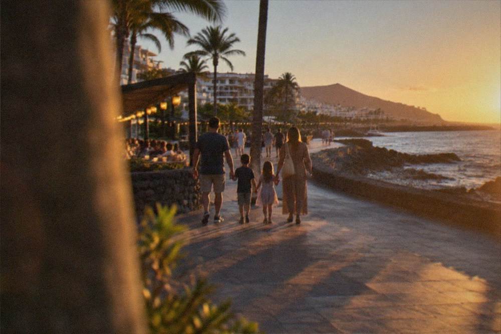 A family walking along a seafront promenade at dusk with lit restaurants behind them