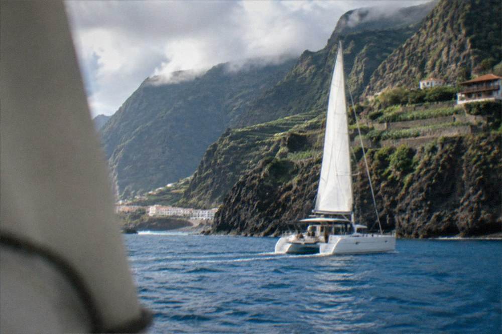 A catamaran sailing close to tall sea cliffs on a bright day
