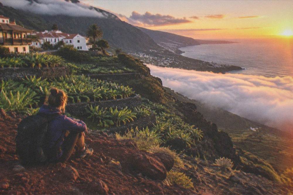 A hiker resting on a red volcanic ridge above a sea of clouds at sunset