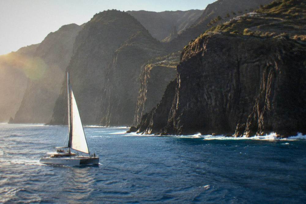 A catamaran under way on open blue water near tall dark cliffs
