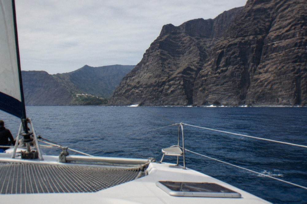 A catamaran under sail near the tall dark cliffs of Los Gigantes on a calm sea