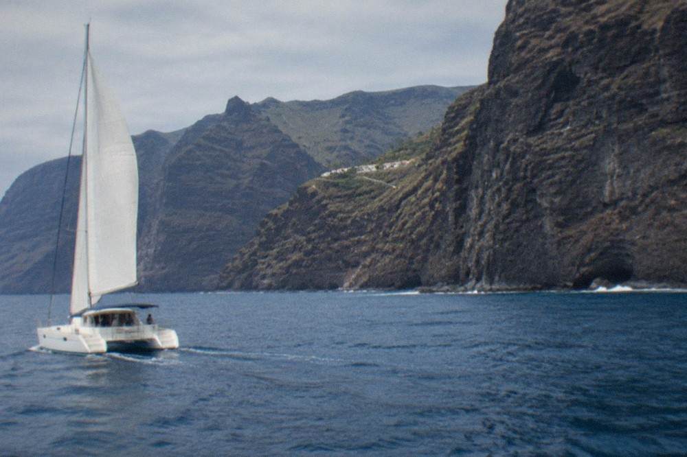 A catamaran under sail near the dark cliffs of Los Gigantes on a calm sea