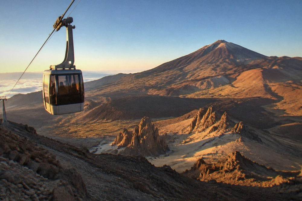 A cable car cabin ascending a rocky volcanic slope above a sea of clouds