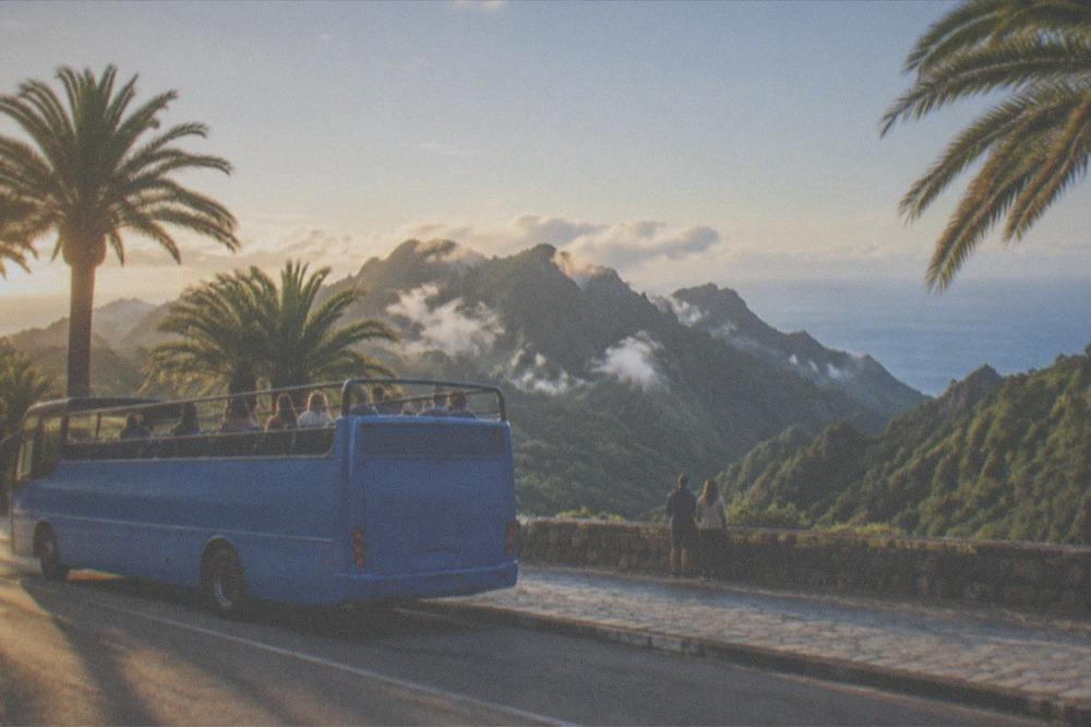 A blue open-top tour bus parked on a sunny seaside promenade with palm trees