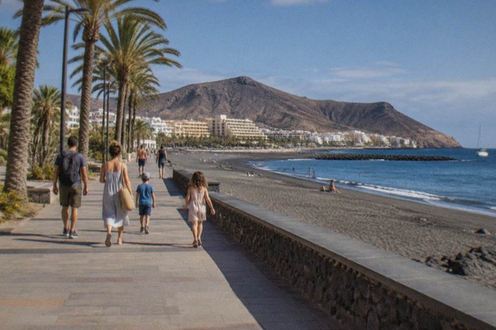 A family walking along a sunny palm-lined promenade beside a calm beach in Tenerife