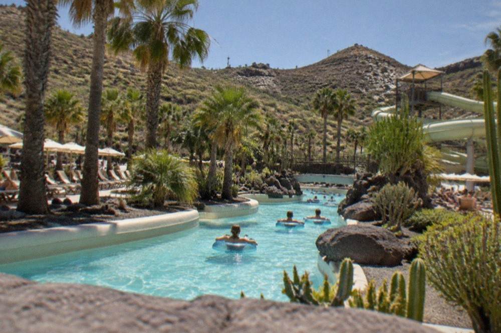 A wide view of a water park with a lazy river winding between palm trees and sunbeds