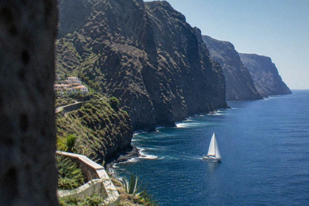 A sailboat passing beneath the tall dark sea cliffs of Los Gigantes on a calm blue sea